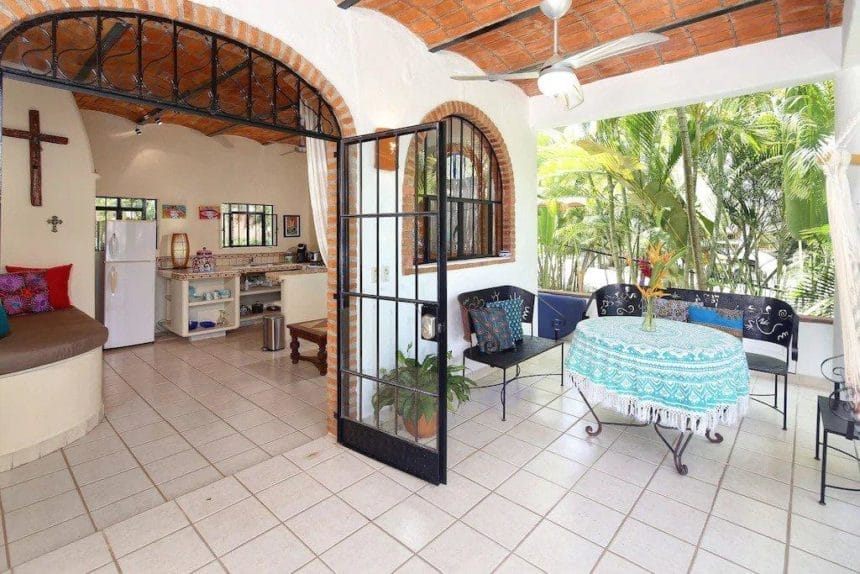 Breezy veranda with arched doorways, wrought iron seating, and a turquoise embroidered tablecloth, overlooking tropical foliage and an airy open-plan kitchen.