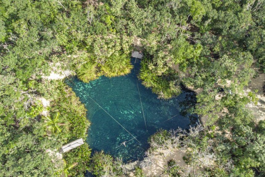 Aerial view of a clear-blue freshwater cenote surrounded by dense green forest. A person can be seen swimming near the center.