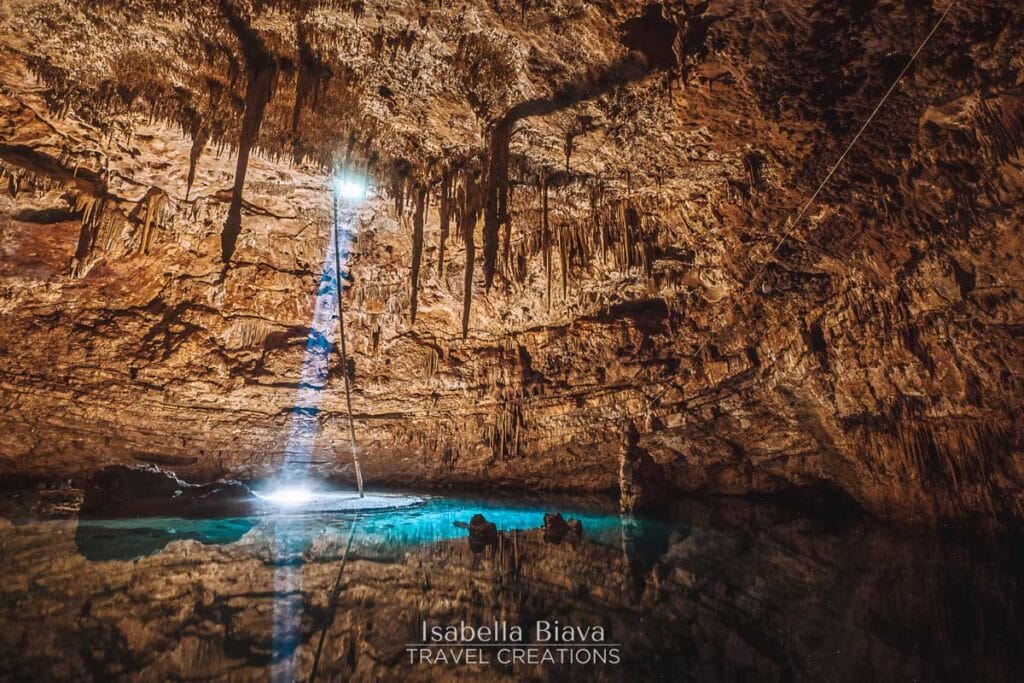 A stunning cave with stalactites and a beam of light illuminating the turquoise water below, creating a mystical atmosphere.