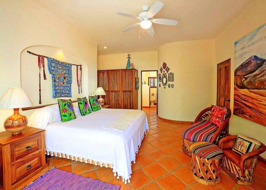 Bright bedroom featuring a king bed with a white coverlet, vibrant embroidered pillows, colorful artwork, and terracotta tiled floors with wood furnishings.