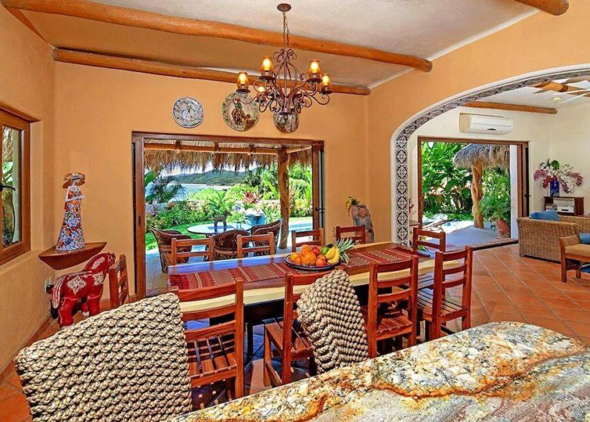 Colorful dining room with a long wooden table, woven-back chairs, a decorative chandelier, and open sliding doors leading to a lush garden and poolside area.