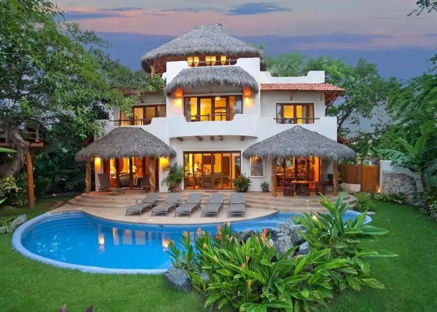 Luxurious multi-level villa surrounded by lush greenery, featuring thatched palapa roofs, glowing interior lights, and a curvy private pool with lounge chairs in the foreground.