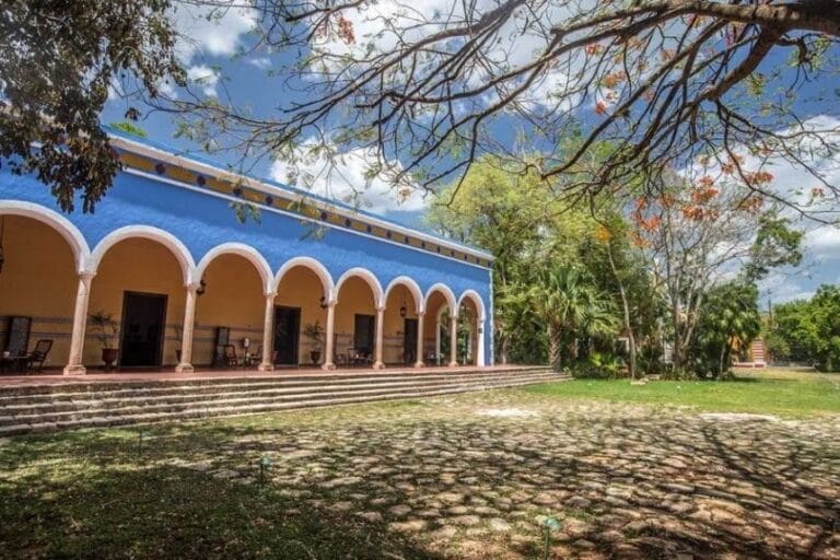 A vibrant blue hacienda with a series of arches stands under a sunny sky, surrounded by lush trees, casting intricate shadows on the ground.