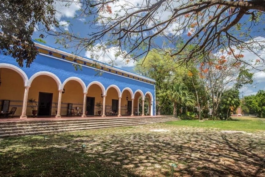 A vibrant blue hacienda with a series of arches stands under a sunny sky, surrounded by lush trees, casting intricate shadows on the ground.