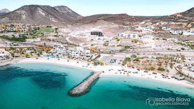 A scenic beach with turquoise water and surrounding hills, featuring white buildings and a mountainous background. Sparse trees and people are visible.