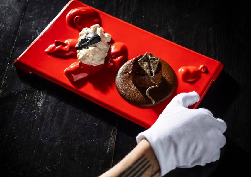 A person with a tattooed arm and gloved hand presents a gourmet dish on a vibrant red platter, featuring sophisticated culinary elements.
