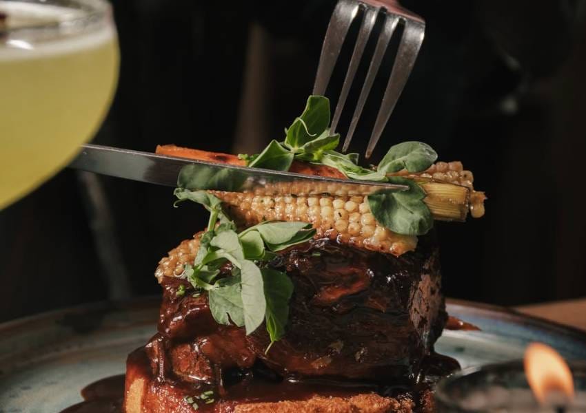 A person cuts a gourmet dish with a knife and fork, featuring roasted meat, greens, and corn, accompanied by a drink and candle.