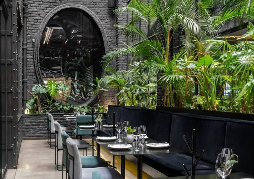 A stylish restaurant interior features black brick walls, lush green plants, elegant table settings, and modern furniture, creating a chic and inviting atmosphere.