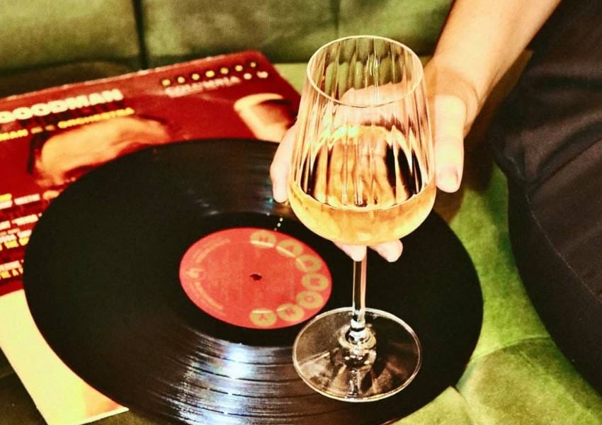 A person holds a wine glass beside a vinyl record and album cover on a green surface. The ambiance suggests a relaxed, vintage vibe.
