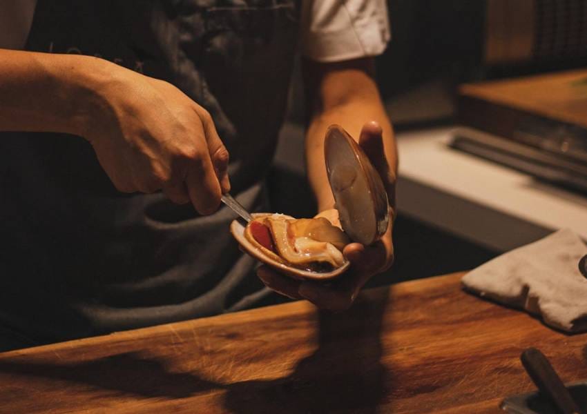 A person expertly opens a clam with a knife on a wooden countertop, preparing seafood in a dimly lit kitchen setting.
