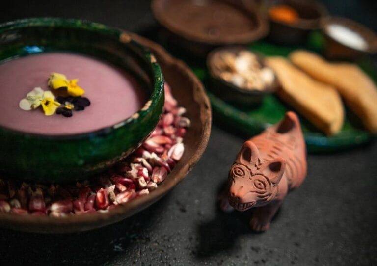 A vibrant, artistic display of food featuring a colorful soup garnished with flowers, surrounded by corn kernels and a small decorative animal figure.