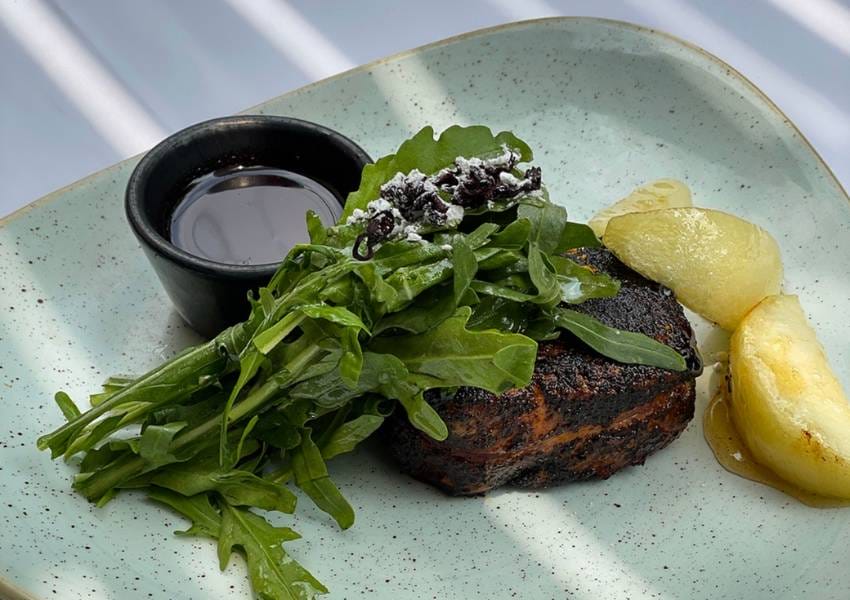 A gourmet dish featuring grilled steak topped with arugula, accompanied by pear slices and a dark sauce in a small bowl, served on a plate.