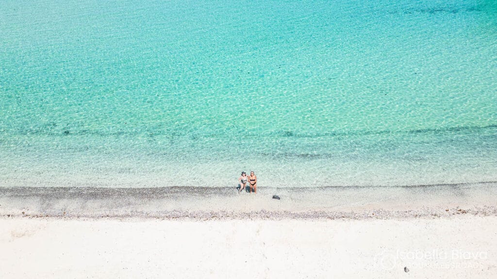 Turquoise water meets sandy beach; two people relax on the shoreline, enjoying the serene scene, surrounded by clear blue ocean.