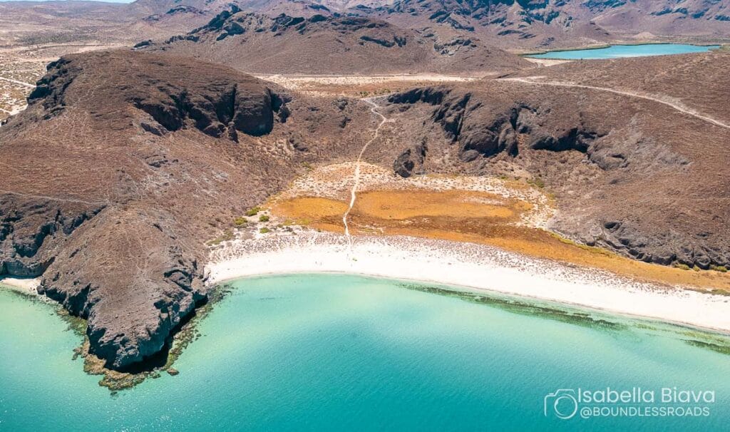 Aerial view of a secluded beach with turquoise waters, surrounded by rugged hills and a narrow path leading inland.