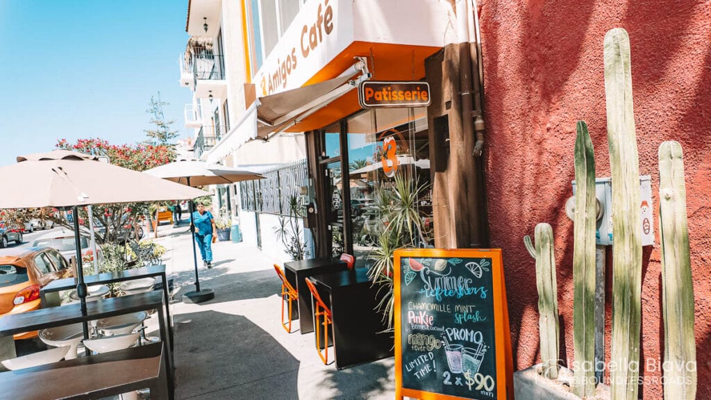 Outdoor café with a colorful sign reading "Patisserie." Person walking by. Tables with umbrellas and cactus plants enhance the lively street scene.