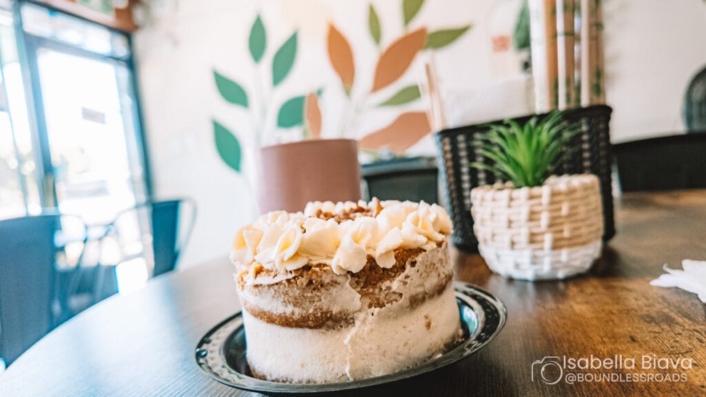 A small cake with frosting is on a table in a cozy cafe, featuring plant decor and a soft, inviting atmosphere.
