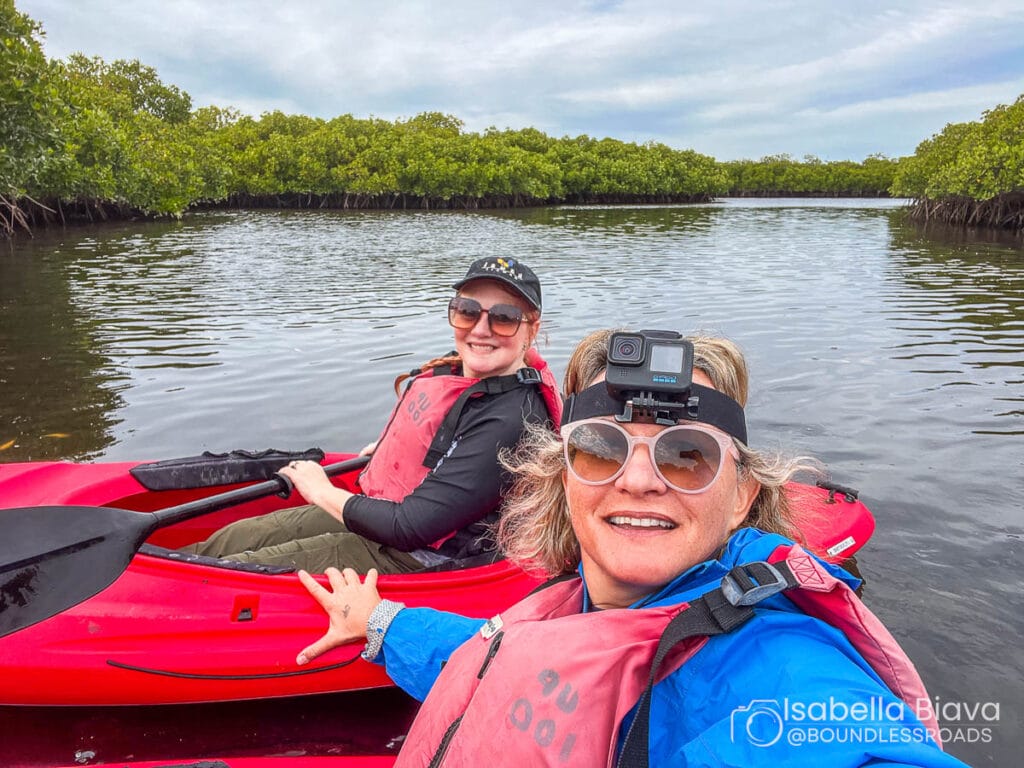 Two people in red kayaks wearing life jackets, explore serene mangrove waters. One person wears a head-mounted camera. Overcast sky in the background.