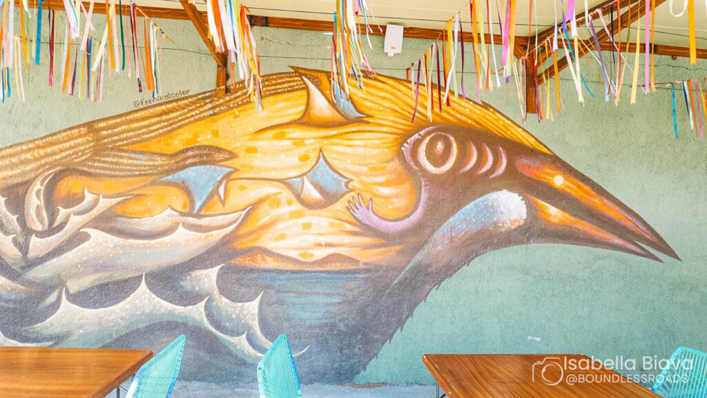 Vibrant mural of a stylized bird adorns a wall, with colorful ribbons hanging above. Wooden tables and turquoise chairs complete the lively scene.
