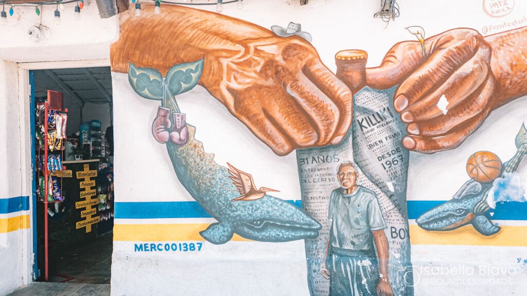 A vibrant mural depicting large hands, a fish, and a person, with a small shop displaying snacks and colorful signage nearby.