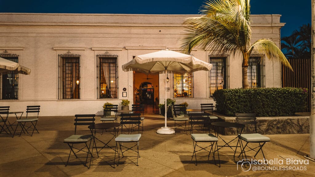 Outdoor café scene at night with empty tables and chairs under umbrellas, set against a historic white building and palm trees.