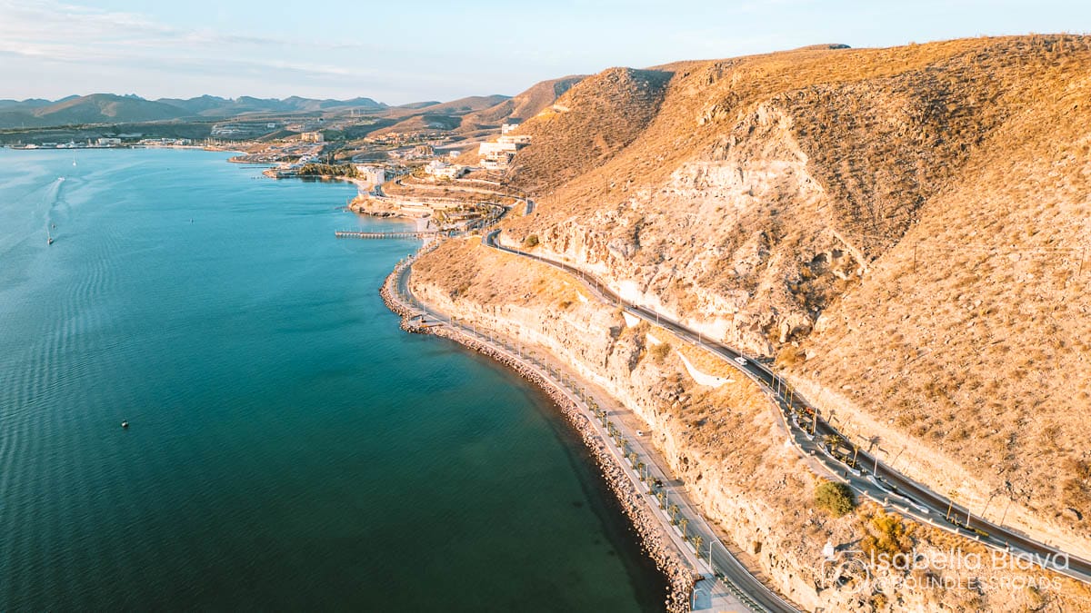 Coastal highway winding along rocky cliffs, overlooking the serene blue waters of the Sea of Cortez. Hills and sparse vegetation surround the area.