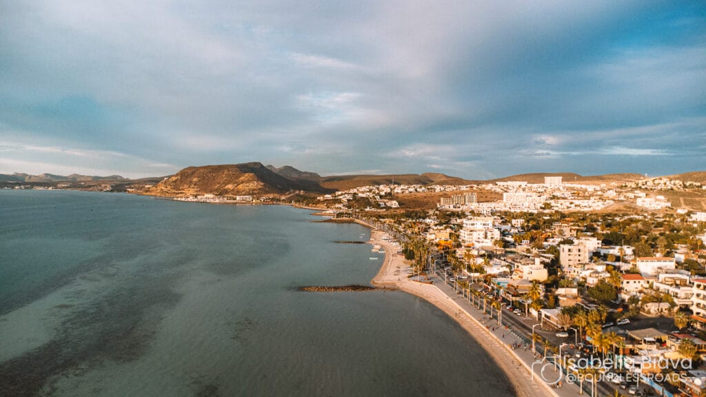 Aerial view of a coastal city with a wide beach, palm-lined promenade, and surrounding hills under a partly cloudy sky.