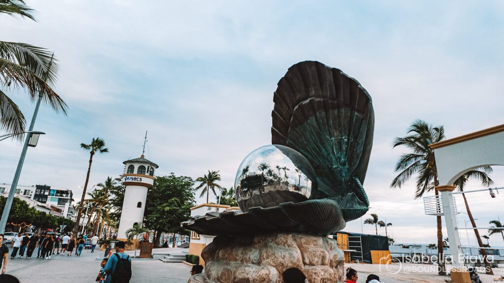 Scenic view of El Malecón in Puerto Vallarta, showcasing a large oyster sculpture and lighthouse, with people walking and palm trees lining the path.
