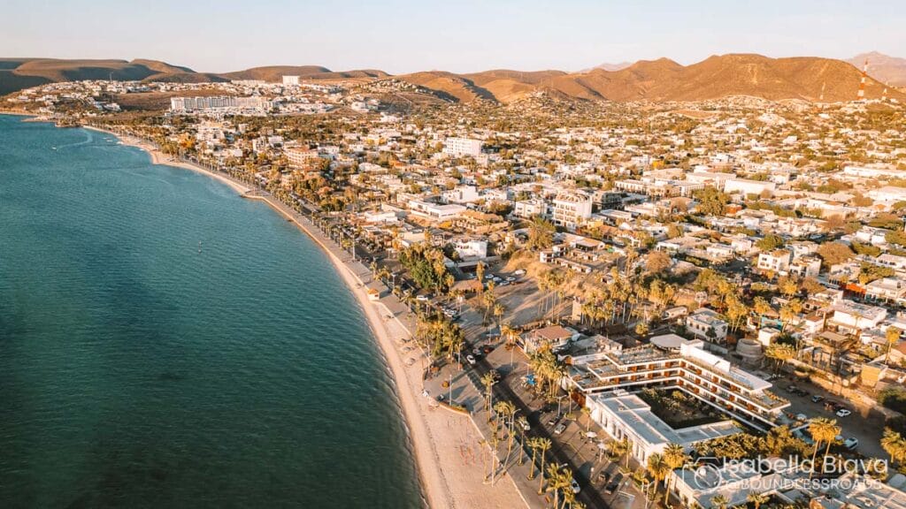 Coastal cityscape features a vibrant beachfront with palm trees. Residential and commercial buildings line the shoreline, set against distant mountains.