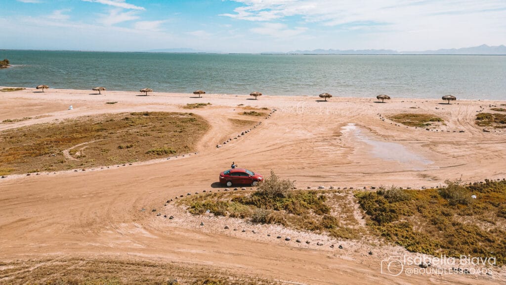 A red car drives along a sandy beach with scattered palapas, overlooking the ocean under a clear blue sky.