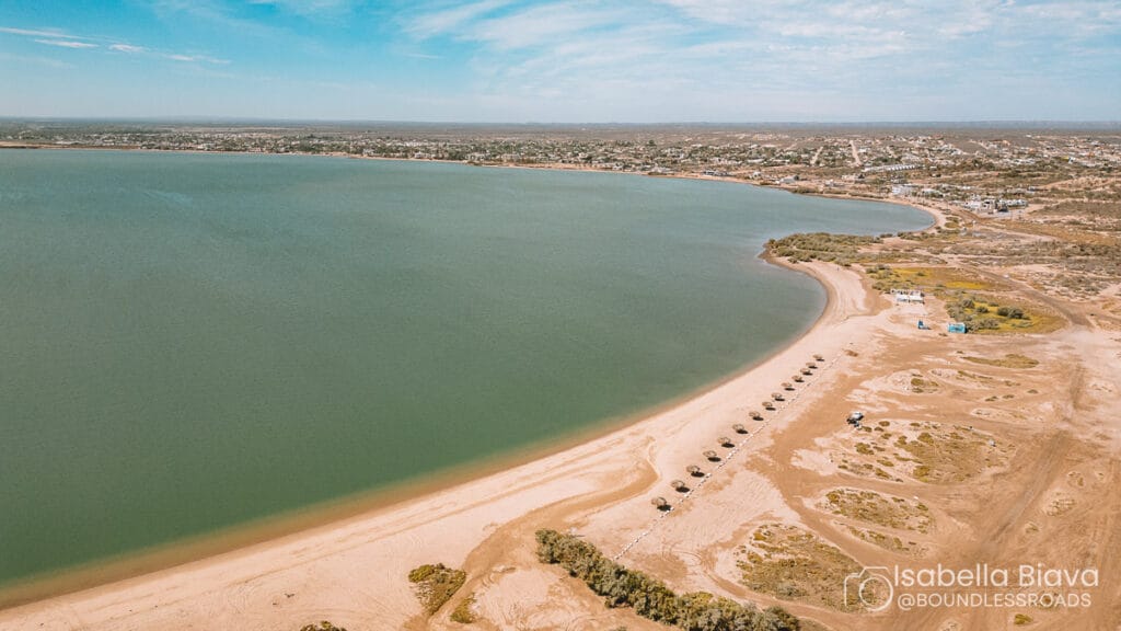 Aerial view of a sandy beach with waterfront umbrellas, calm waters, and a distant town under a clear blue sky.