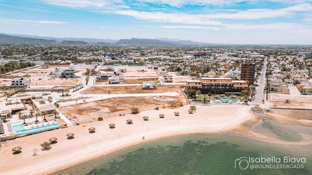 Aerial view of coastal cityscape featuring a beach with sunshades, residential areas, and mountains in the distance under a clear blue sky.