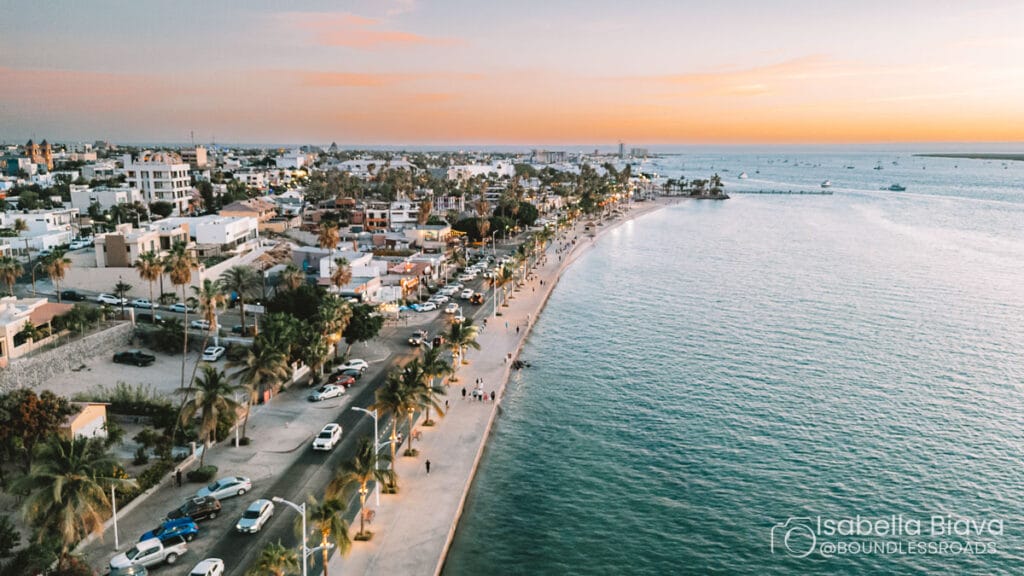 A coastal cityscape at sunset with cars, palm-lined promenade, and people strolling, bordered by ocean waves reflecting warm, colorful skies.