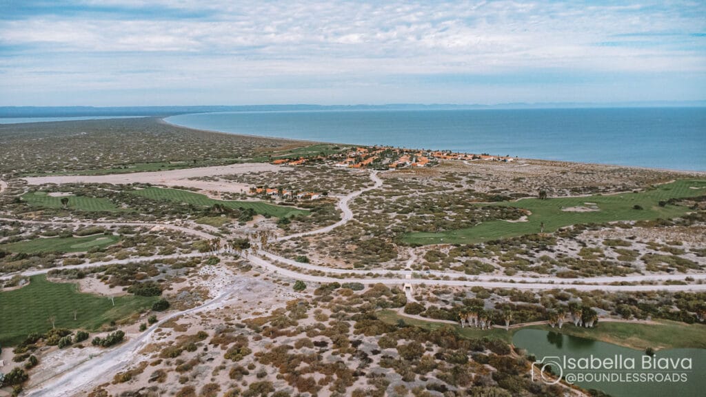 Aerial view of Todos Santos with lush golf course, desert terrain, and coastline, leading to a distant horizon under a partly cloudy sky.