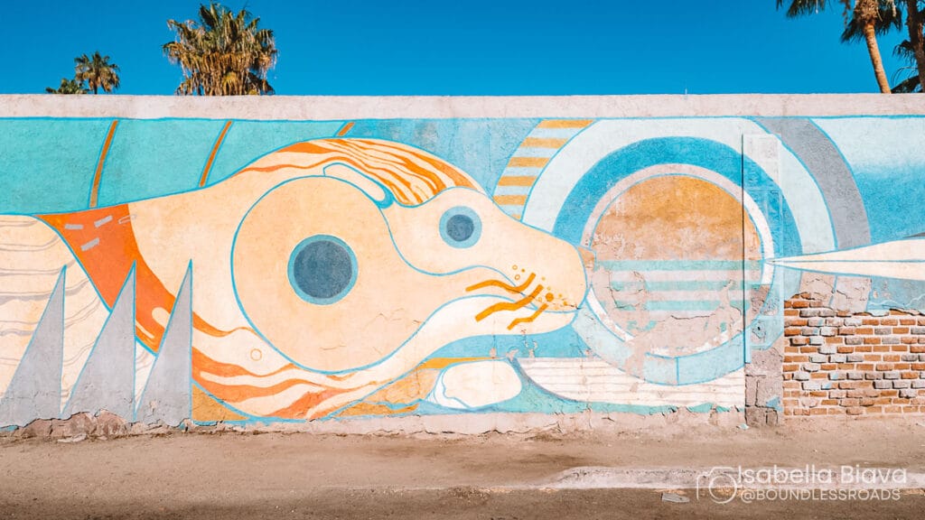 Colorful mural depicting abstract animal shapes and circles on a wall, under a clear blue sky with palm trees in the background.