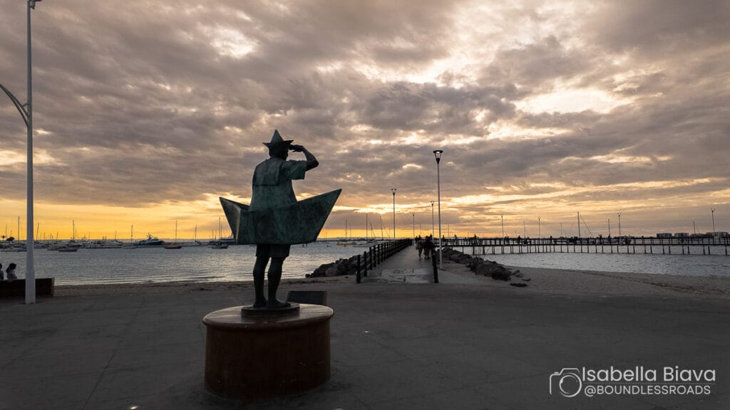 A statue depicting a person in a paper boat overlooks a sunset at the waterfront, with sailboats and a long pier visible.