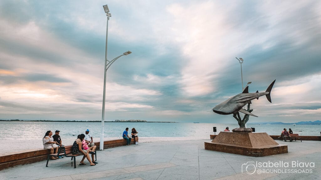 People sitting near a shark sculpture by the ocean under a cloudy sky. The scene captures a peaceful coastal atmosphere.