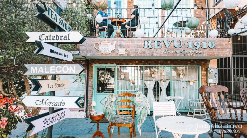 Quaint café with outdoor seating, colorful signage pointing to landmarks like "Catedral" and "Malecón," and two people dining on the balcony.