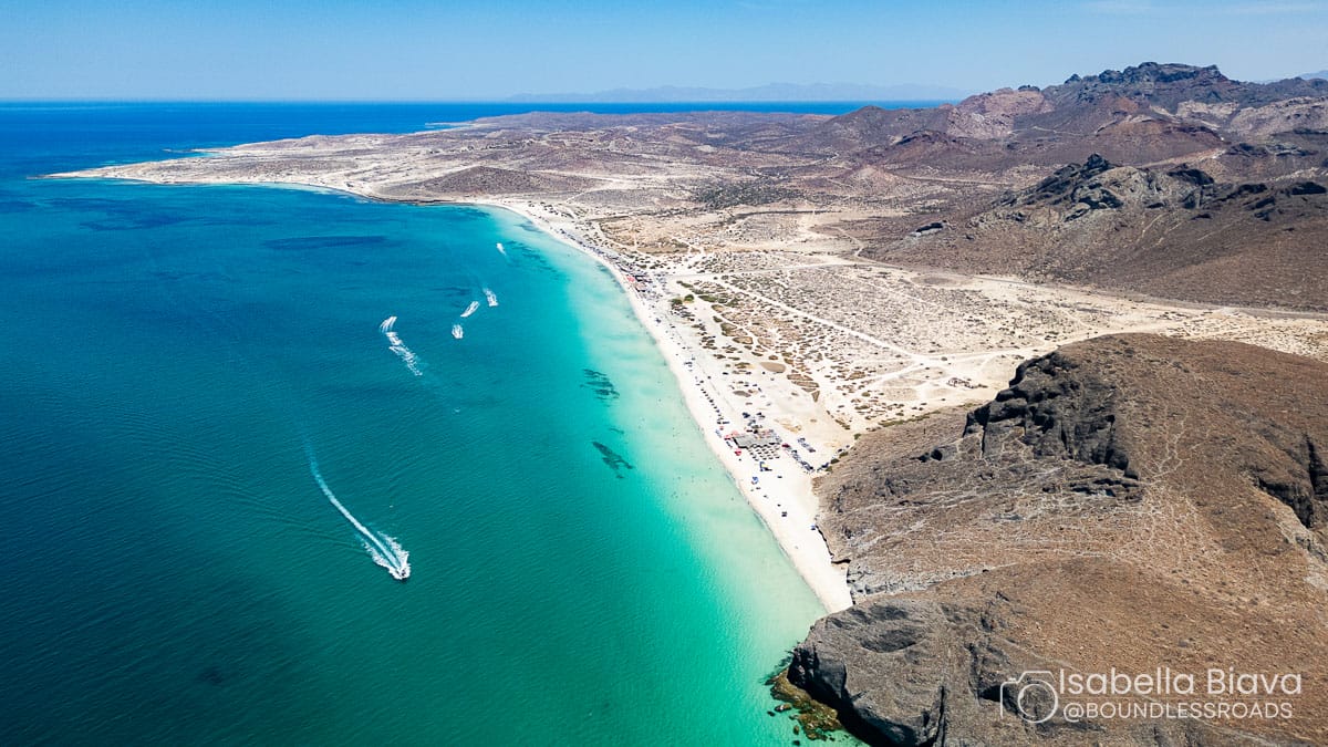 Aerial view of Playa Balandra's turquoise waters, sandy shore, rocky cliffs, and boats. Desert landscape surrounds the serene coastline under clear skies.