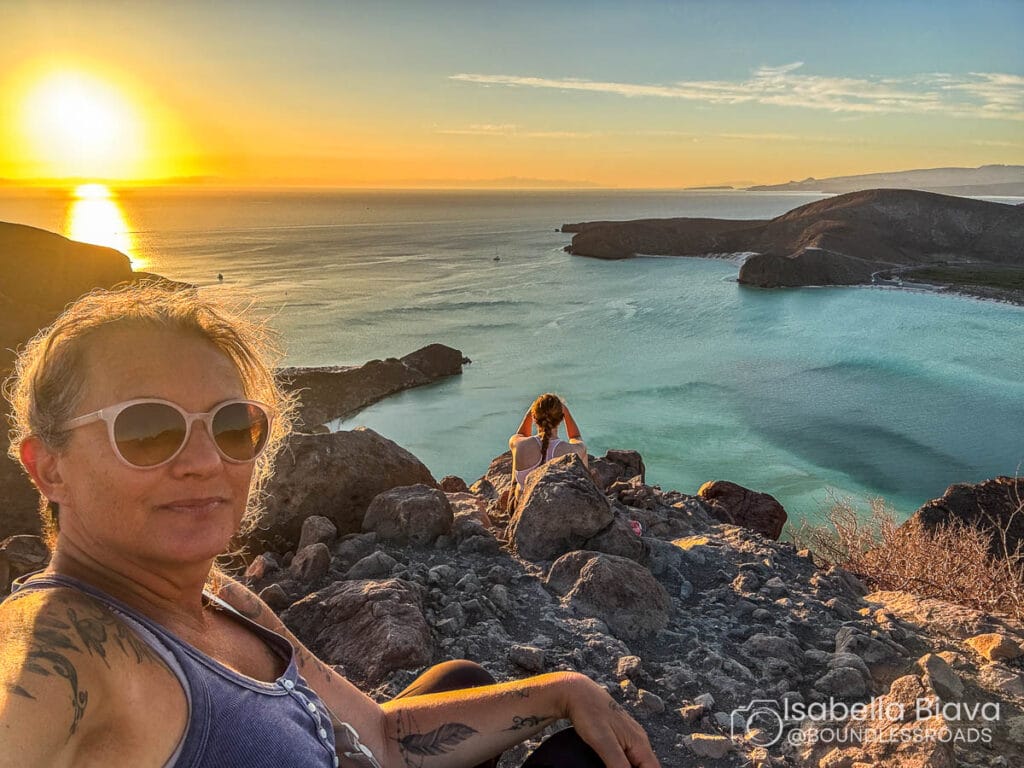 A person enjoys sunset atop a rocky cliff overlooking a calm ocean and distant hills, with another person sitting nearby.