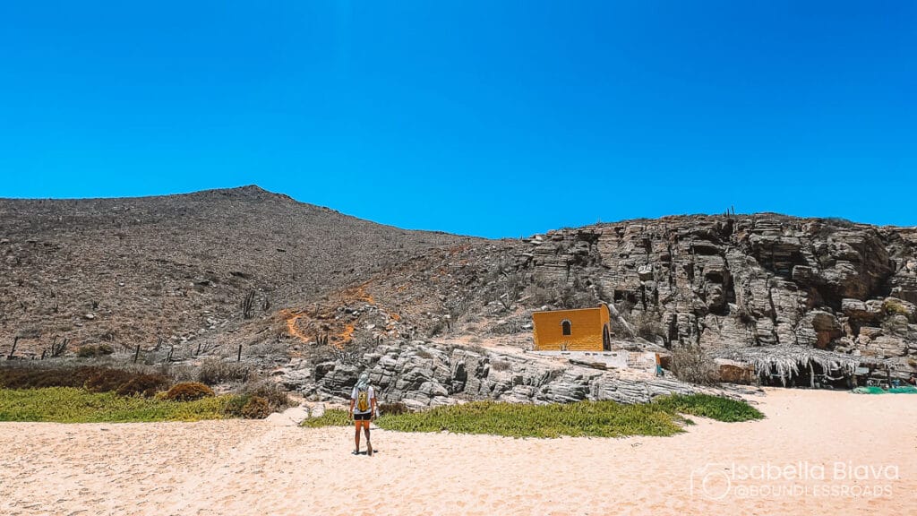 A person approaches a small yellow chapel surrounded by rocky desert terrain under a clear blue sky.