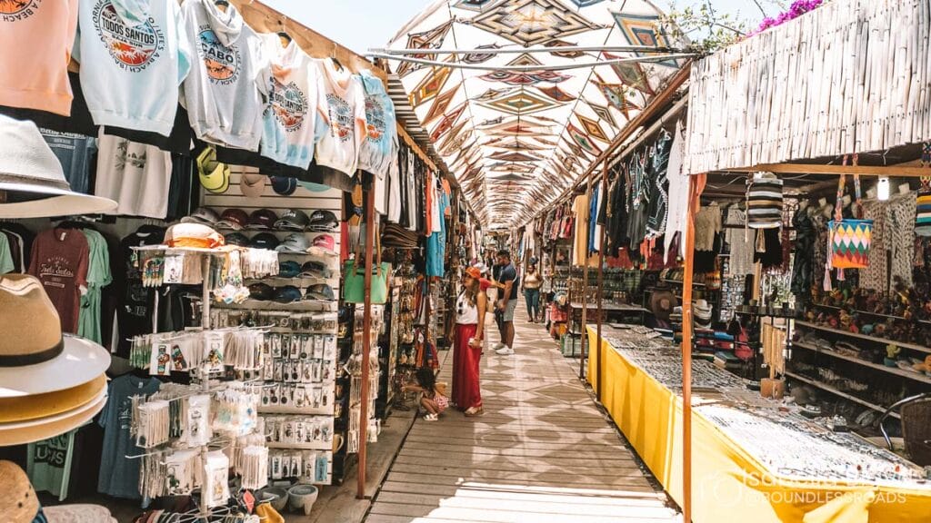 Bustling outdoor market with colorful clothing and souvenirs on display. Several people browse stalls beneath decorative overhead canopy on a sunny day.