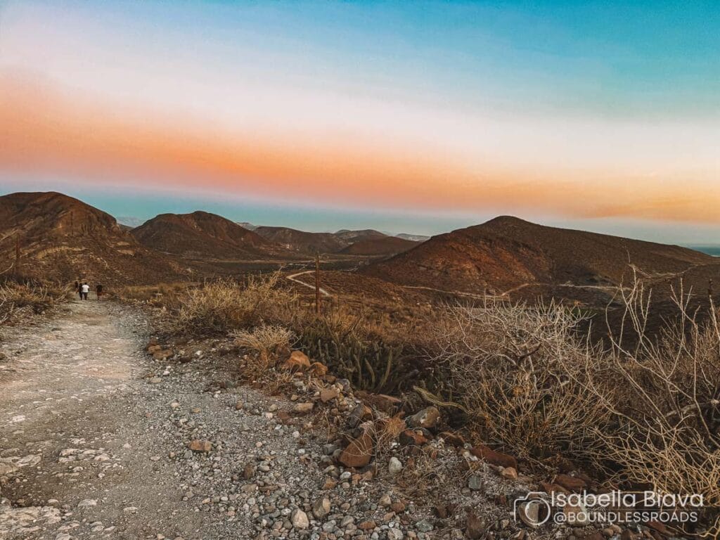 A rocky path winds through a desert landscape at sunset, with two people walking toward distant mountains under a colorful sky.