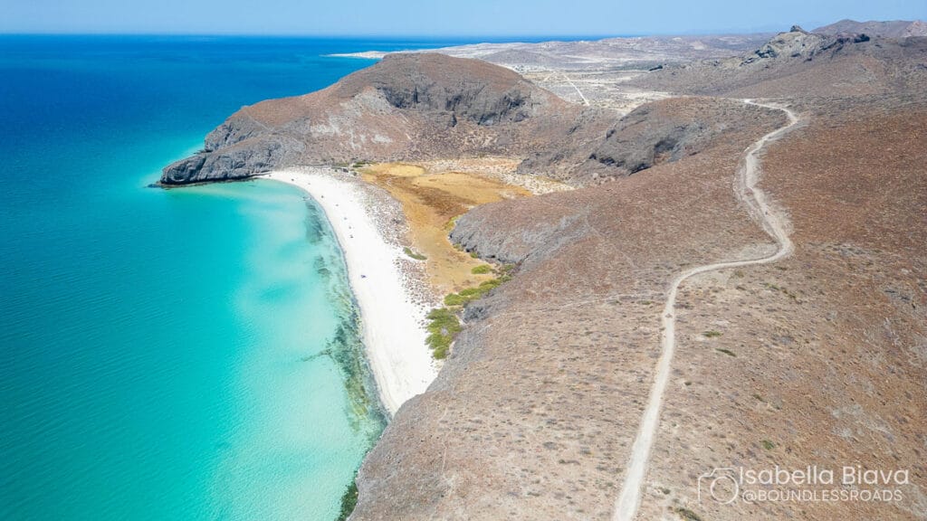 A secluded beach with clear turquoise waters, surrounded by rugged hills and a winding dirt path, against a vast, distant landscape.