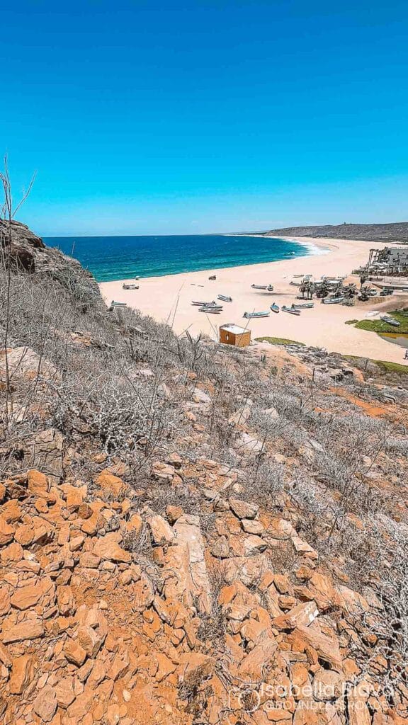 Rugged landscape overlooking a deserted beach with several small boats. Blue ocean meets sandy shoreline under clear sky. No people visible.