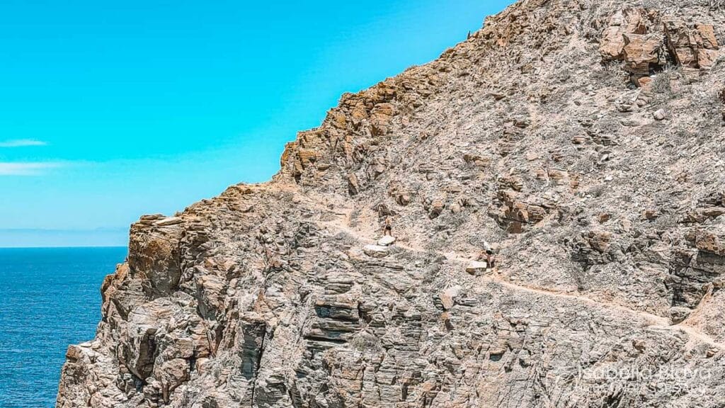 A rocky cliffside with two people hiking along a narrow trail overlooks a vast ocean under a clear blue sky.