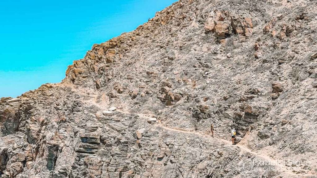 A person hikes along a narrow, rocky mountain trail under a clear blue sky. The terrain is barren and rugged.