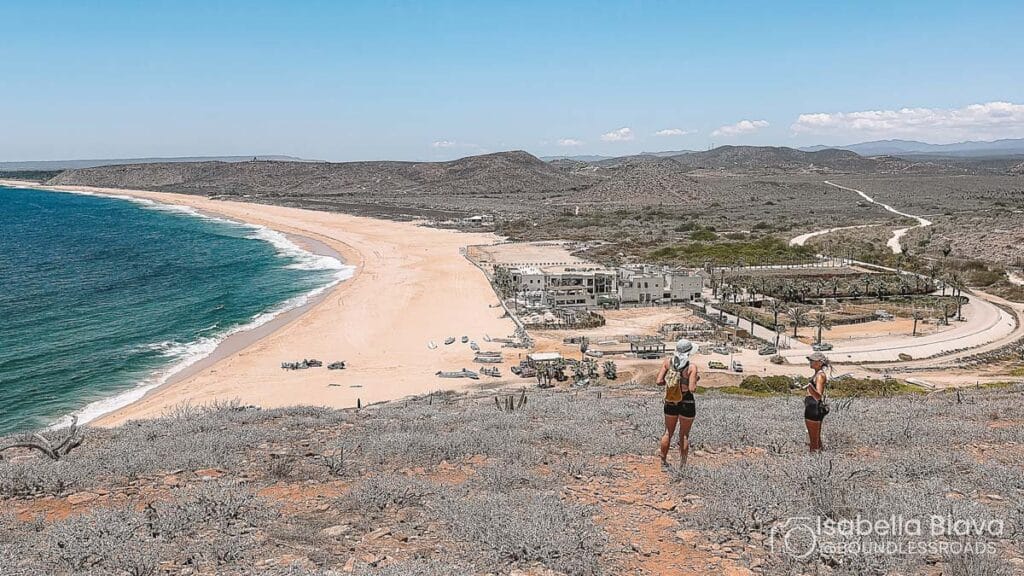 Two people stand on a hill overlooking a vast, empty beach with waves, buildings, and rugged terrain in the background.