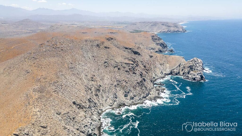 Aerial view of the rugged, rocky coastline of Cabo Pulmo, with deep blue ocean waves crashing against the cliffs under a clear sky.