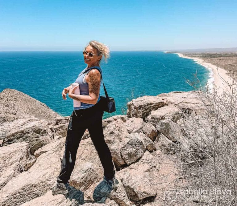 A person stands on rocky cliffs overlooking the ocean, with a vast, clear blue sky and coastline in the background.