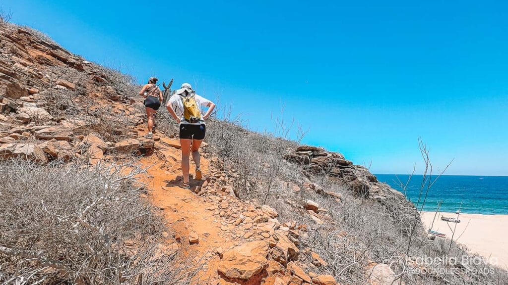 Two people hiking up a rocky, dry trail along a coastal landscape with clear blue skies and ocean in the background.